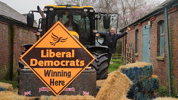 Ed Davey waves from a tractor with a Lib Dem Diamond sign, having knocked down a blue wall of bales